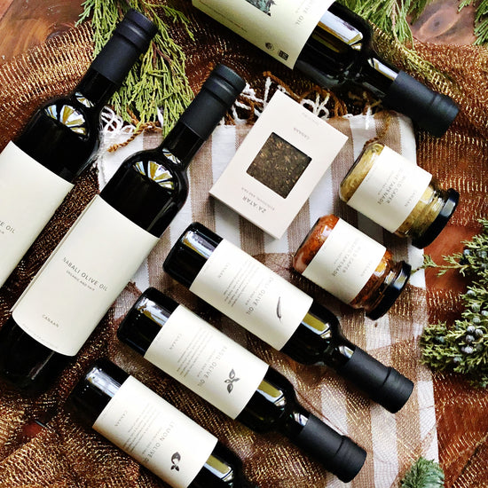 Wine bottles, placed on wooden furniture, surrounded by cypress plants, olive oil bottles, olive oil tapenade, and wild caper tapenade