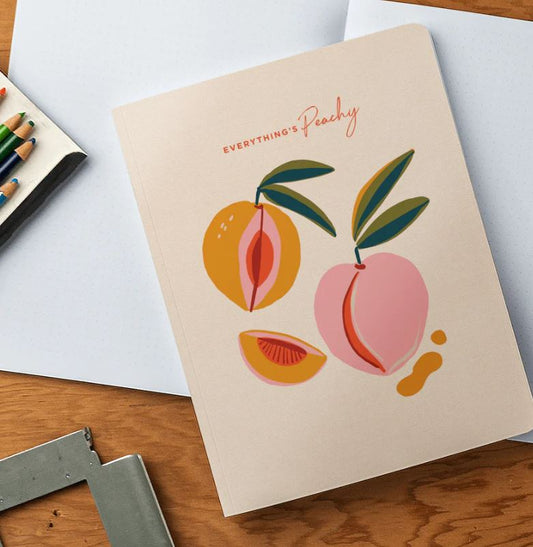 A notebook adorned with a drawing of peaches rests atop an open notebook with two blank pages, accompanied by a colorful pencil case, all on a warm wood surface